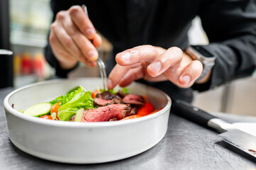 A close-up of hands garnishing a dish with fresh green lettuce, featuring sliced red meat and colorful vegetables in a white bowl. Perfect for culinary, food preparation, and gourmet dining themes.