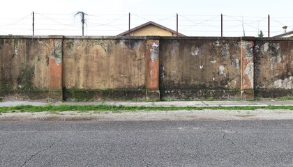 Worn grunge surrounding wall with barbed wire on top. Weathered sidewalk, weeds and road in front....