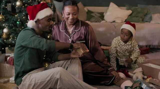 Modern African American family with kid wearing pyjamas opening Christmas gifts while sitting on floor in cozy living room