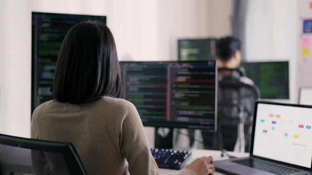 A female developer is observed from behind, scrutinizing a complex codebase on her monitor within a modern technological workspace, indicative of her problem-solving capabilities.