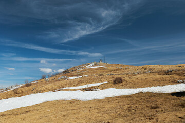 ampia vista panoramica della cima e parte dei pendii erbosi e secchi del Monte Matajur, coperto parzialmente dalla neve, sotto un cielo azzurro, di mattina, in primavera