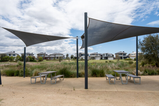 Outdoor recreational area featuring modern shade sails over picnic tables in a suburban neighborhood with contemporary residential houses. Triangular shade on metal poles provide protection from sun.