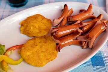 piatto con pezzi di wurstel accompagnati da una composizione di patate di forma rotonda,...