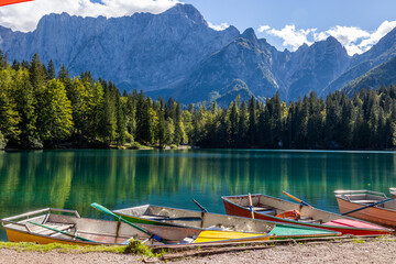 vista dettagliata del lago di Fusine inferiore visto da una delle sponde, con alcune barche ormeggiate e la distante catena montuosa del monte Mangart, in estate