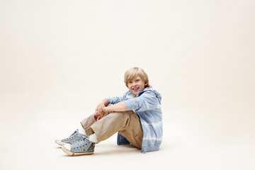 Studio shot of cheerful preteen boy wearing blue sneakers and zip hoodie sitting on floor while smiling at camera against neutral background in studio, copy space