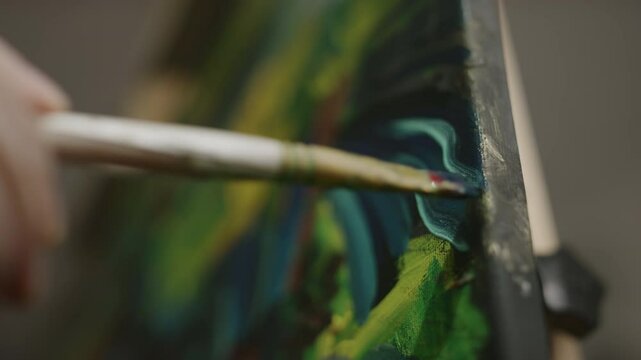 Close-up shot of hand of anonymous artist adding fine details in turquoise colour to abstract oil or acrylic painting, while working on easel in workshop