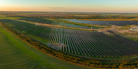Rows of solar panels in a Lithuanian solar farm stretch across a flat, rural landscape. The setting sun casts a warm glow over the organized installation.