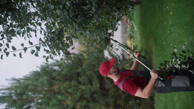 Gardener in uniform cuts tree branches using a telescopic lopper in a well-maintained garden. The scene captures the skill and precision involved in professional landscaping work
