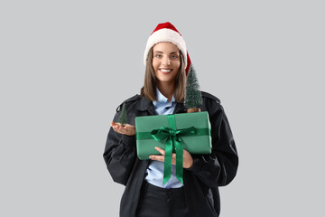 Female police officer in Santa hat with toy Christmas trees and gift on light background