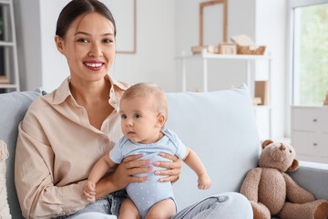 Beautiful young happy Asian mother with her cute little baby sitting on sofa at home