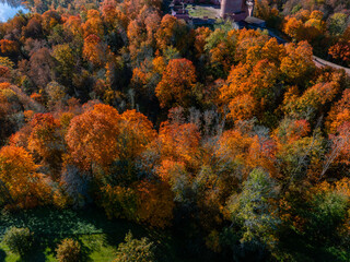 An aerial view of Turaida Castle in Sigulda, Latvia, surrounded by vibrant autumn foliage. The red brick tower contrasts with the colorful forest and blue sky.
