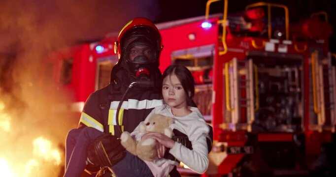 Portrait of young African American brave firefighter standing at night outdoor and holding small cute saved girl with teddy bear on hands. Saving child from fire. Red water truck on background.