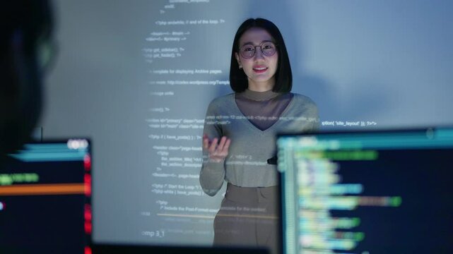 A female developer conducts a coding workshop at night, with HTML code projected on a screen in an office collaboration setting.