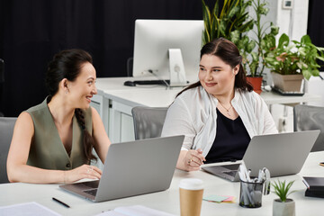 Two professional women engage in a lively discussion while working on their laptops in the office.