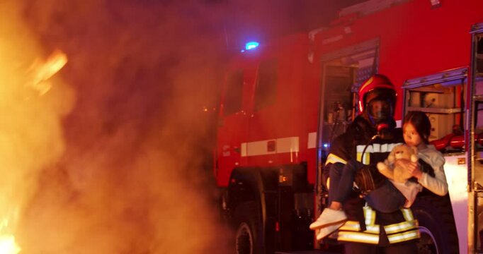 Brave full equipped fireman holding little girl with teddy bear and taking her out from fire outdoors. Open fire and smoke. Red water truck on background. Courageous fireman saving life small child.