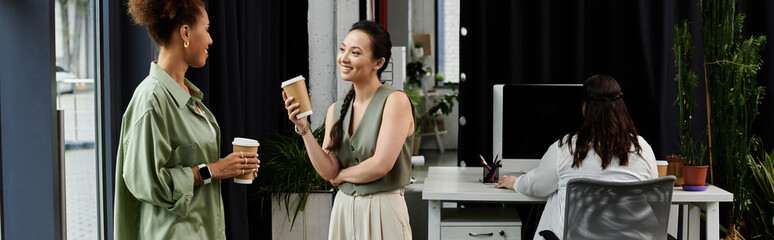 Two stylish businesswomen chat leisurely while holding coffee cups in a chic office space.