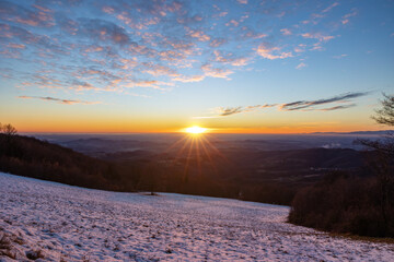 sole a filo sull'orizzonte che tramonta e colora il panorama e l'ambiente naturale di sfumature di arancione, visto dall'alto lungo un pendio innevato tra collina e pianura tra Slovenia ed Italia