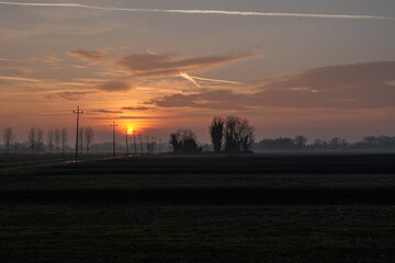 sole che tramonta all'orizzonte da un ambiente di campagna in pianura, nel nord est Italia, con cielo nuvoloso, in autunno