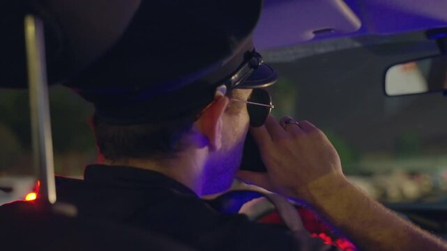Police officer in patrol car at night calling on radio to dispatch or police station. Waiting for backup or reporting on crime or suspicious activity outside in city street. Male cop inside vehicle