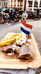 Traditional Dutch herring served with pickles and onions, topped with a Netherlands flag. Captured in a vibrant Amsterdam street, this image showcases local flavors and culture in a deliciously iconic