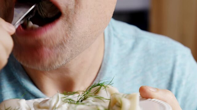 man with Fork takes and eats dumplings from the plate. Boiled dumplings with dill and cream are arranged in a circle on a white plate.