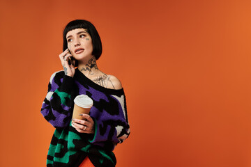 A young woman with short hair and tattoos poses confidently in a colorful sweater while holding a coffee.