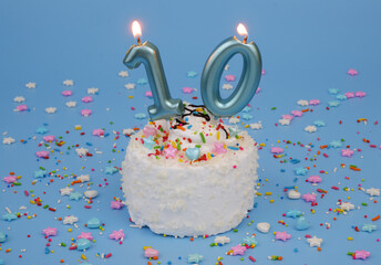 Coconut birthday cake on blue background with burning candles number 10 and many colorful sprinkles.