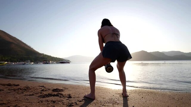 A woman in a sports pose with dumbbells on the beach, her muscles are working, and the surrounding nature and the view of the mountains enhance the feeling of harmony and strength.