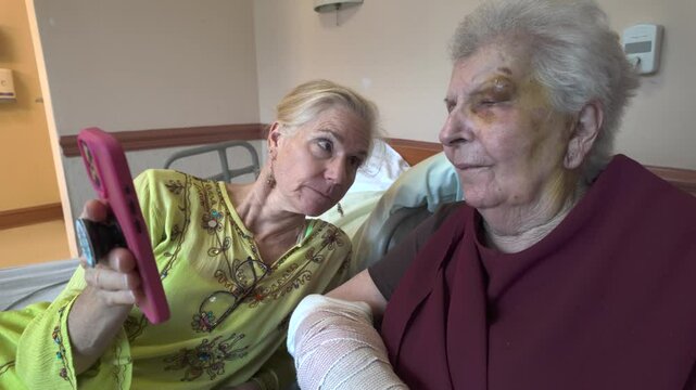 A young woman holds a pink smartphone with a pop socket, taking a picture of an elderly woman who has a bandaged arm and is sitting in a hospital bed. 