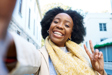 Joyful Woman Taking a Selfie Outdoors in a City Street, Showcasing a Peace Sign and a Bright Smile