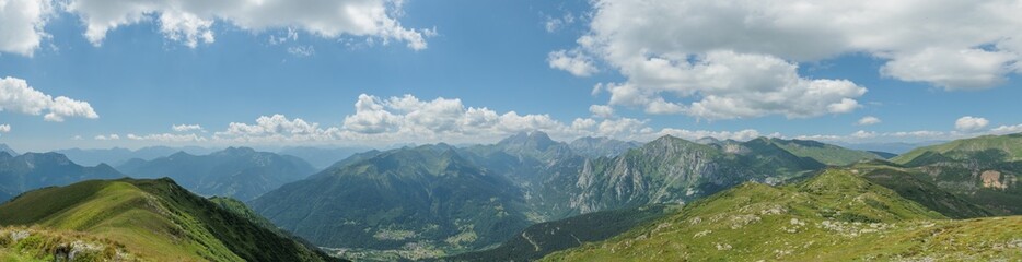 panoramica ultra-ampia su delle catene montuose erbose nelle Alpi Italiane, di giorno, tra primavera ed estate, sotto un cielo azzurro, coperto da qualche nuvola