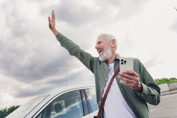 Photo of handsome good mood seniour guy wear green shirt rising arm catching car taxi modern gadget outdoors urban city street © deagreez