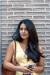 Young latin woman is standing in front of a brick wall, holding her smartphone and looking at the camera