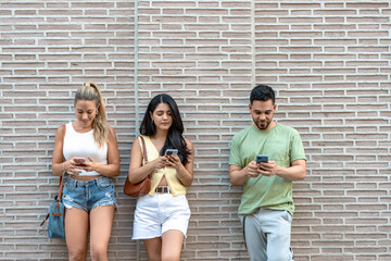 Three friends standing against a brick wall in the city, engrossed in smartphones, browsing, scrolling, and texting, showcasing the modern lifestyle of constant digital connection