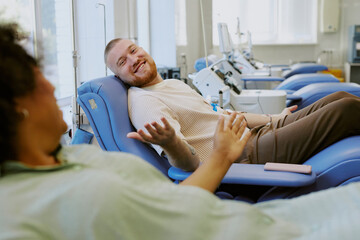 Smiling man lying on medical chair donating blood, engaging in conversation with person beside him. Medical equipment visible in background providing a clean, professional environment