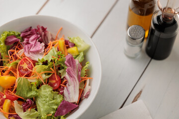 grande scodella bianca piena di verdure miste e di vari colori su una superficie di legno bianco, visto da sopra, con una salvietta e vari recipienti per condire con sale, aceto ed olio