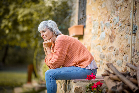 Senior woman sitting outdoors in a thoughtful pose near a rustic home