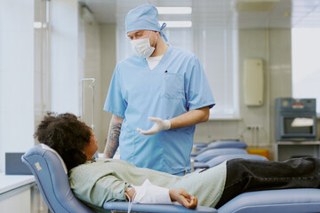 Fototapeta premium Medical professional in blue uniform assisting African American patient in clinic. Patient lying on reclining chair receiving medical attention from professional