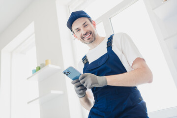 Smiling handyman in blue overalls using smartphone during home renovation in bright modern apartment
