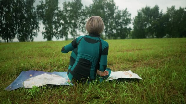 Back view of an older woman seated on a colorful mat in siddhasana pose, practicing yoga in a vast grassy field under a cloudy sky, with trees lined up in the background