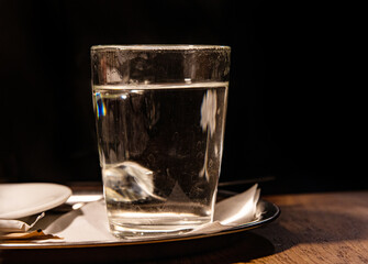 Close up of glass with hot water served on the metal tray with white napkin, sugar pack and tee bag against black background. Transparent tea cup on the café table. Copy space
