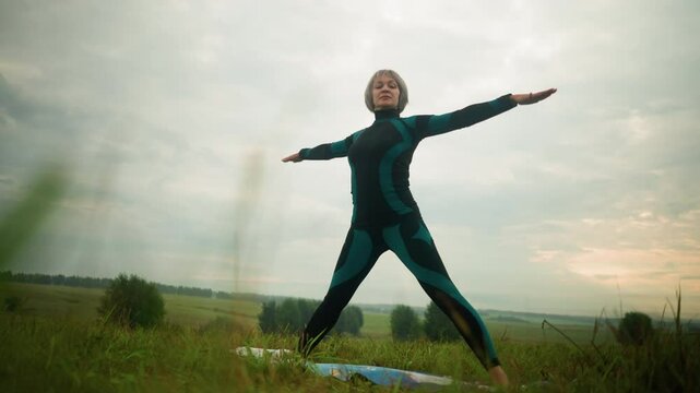 Low-angle view of woman in green-black suit standing on yoga mat practicing yoga triangle pose with arms open and legs apart, set against cloudy skies in a serene, grassy field