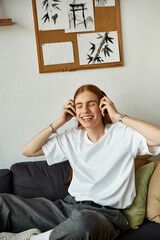 A young man relaxes on a couch, wearing headphones and smiling as he listens to music.