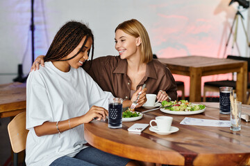 Two women share a loving moment at a cafe, savoring fresh salads and drinks while smiling warmly.