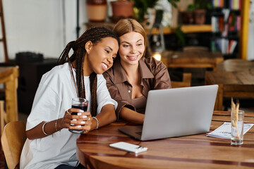 Two joyful women sit together, enjoying each others company and a laptop, surrounded by a vibrant cafe atmosphere.