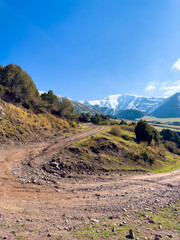 A dirt road winding through the mountain slopes in Kyrgyzstan, with snow-capped peaks in the background and a clear blue sky.