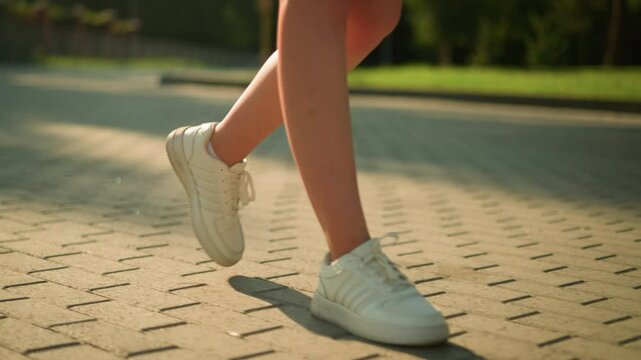 Leg view of white lady wearing white canvas shoes walking along interlocked path, shadow cast on ground, with blur background featuring greenery and soft light glow