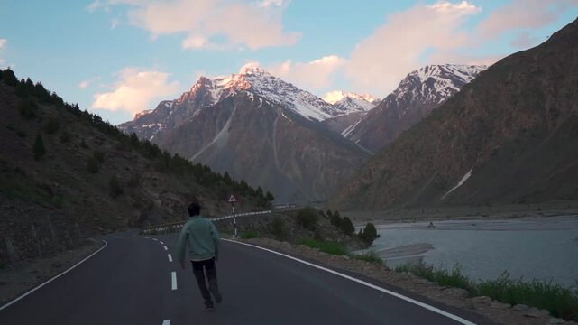 Slow motion shot of an Indian man running on the road in the Himalayan mountain peaks during the sunset at Jispa in Lahaul Valley, Himachal Pradesh, India. Tourist runs in front of snowy Himalayas.