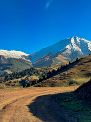 A dirt road winding through the mountain slopes in Kyrgyzstan, with snow-capped peaks in the background and a clear blue sky.