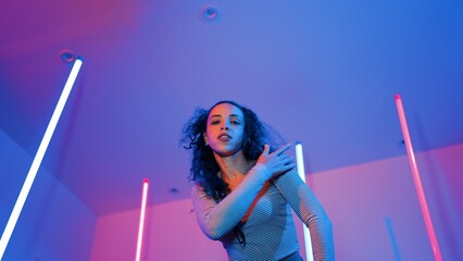 Low angle camera of hispanic dancer looking at camera and dancing to city pop music. Professional attractive hipster moving to hip hop song with purple led or neon light. Stylish cloth. Regalement.